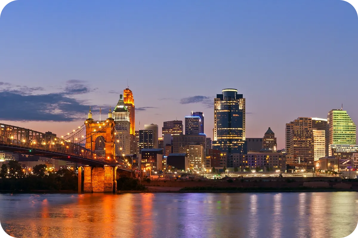 City skyline at dusk with illuminated bridge and buildings reflecting on a calm river under a clear sky.