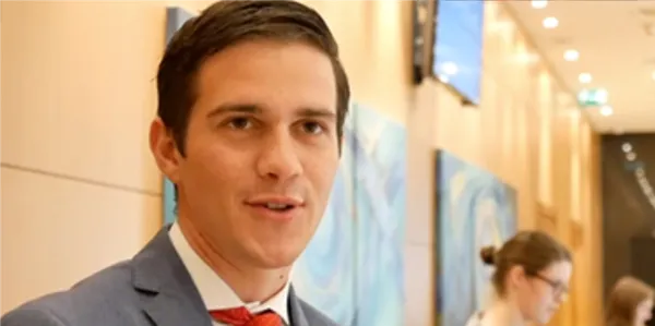 A man in a suit and red tie is speaking indoors, with abstract artwork and another person blurred in the background.