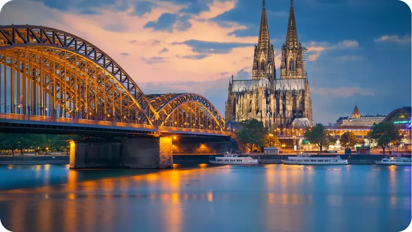 Cologne Cathedral illuminated at dusk beside the Hohenzollern Bridge over the Rhine river.