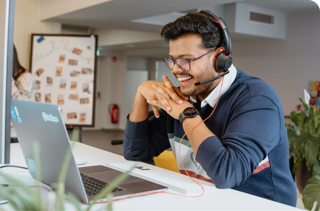 Man wearing glasses and a headset, smiling at a laptop in a bright office space with plants and a whiteboard in the background.