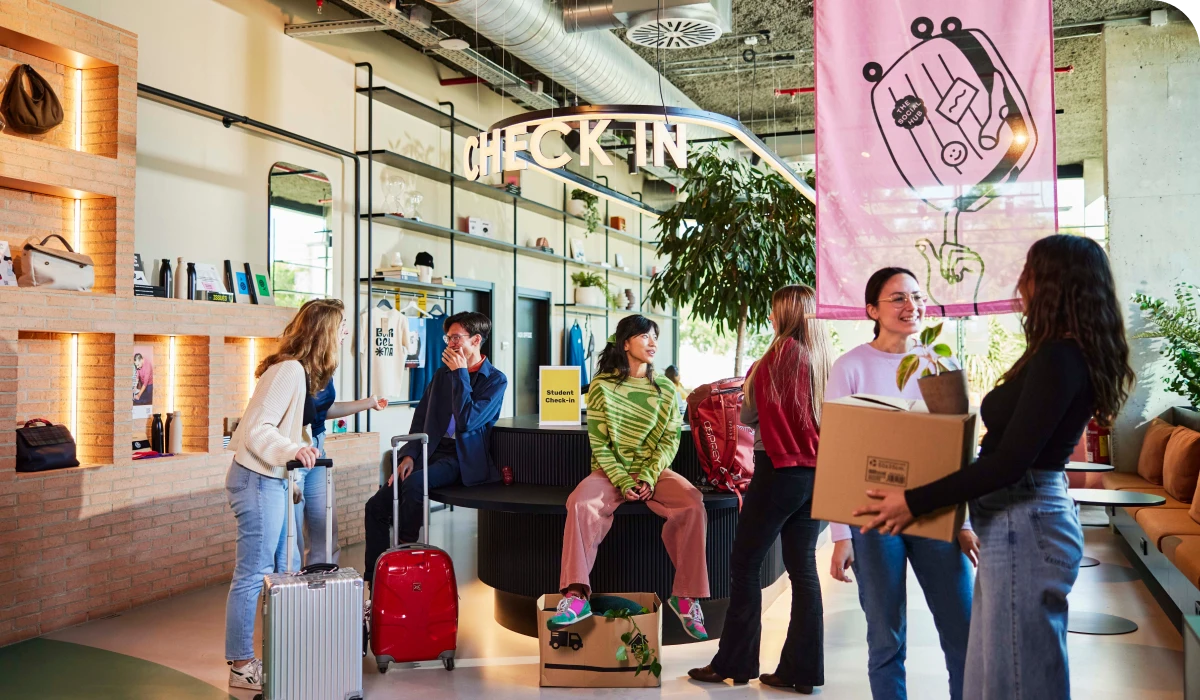 People with luggage and boxes gather around a modern, brightly lit hotel check-in area with plants and a pink banner.