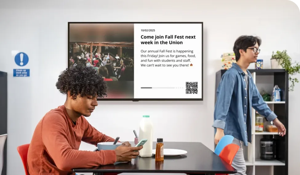 A person sits at a table with milk and snacks, using a phone. Another person walks by. A screen displays a Fall Fest announcement.