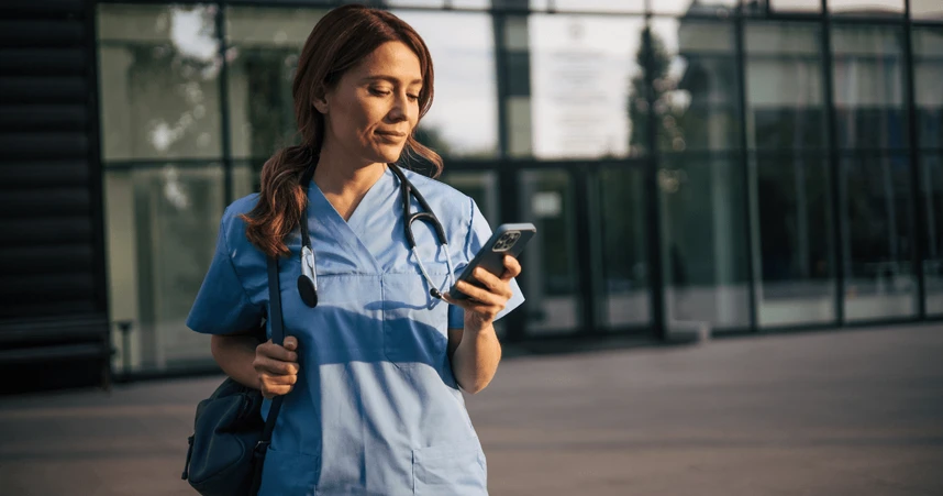 A healthcare professional in blue scrubs and a stethoscope checks her phone while standing outside a modern building.