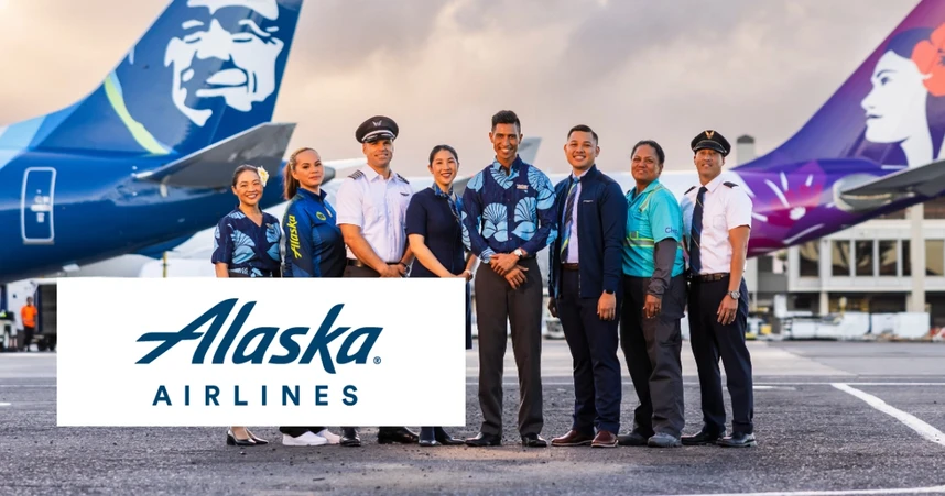 Alaska Airlines crew in uniform stand smiling on the tarmac, with two airplanes in the background and the Alaska Airlines logo in the foreground.