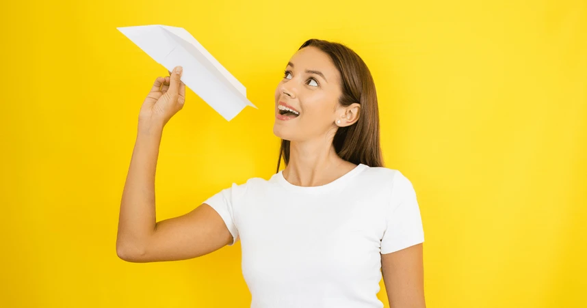 Smiling woman in a white t-shirt holding a paper airplane, preparing to throw it, against a bright yellow background.