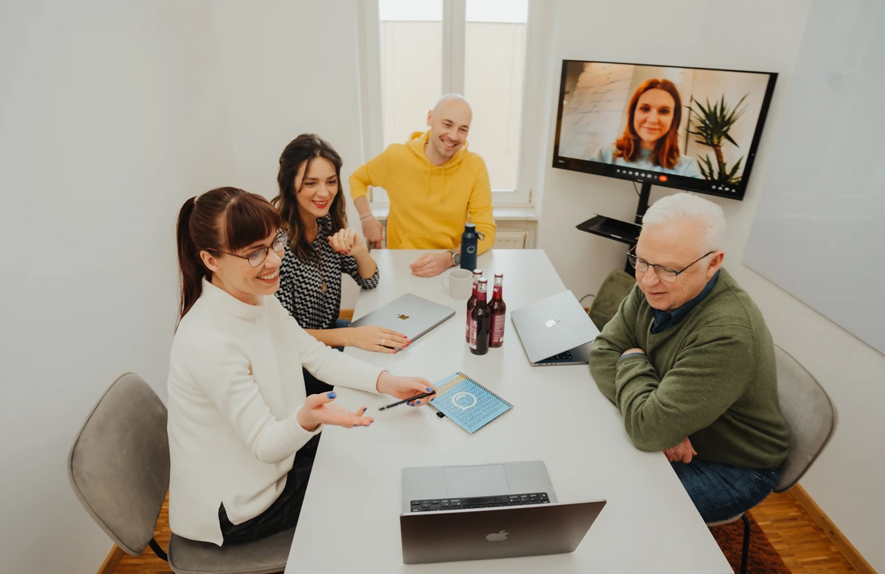 Four people in a meeting room with laptops and drinks, video conferencing with a person on a screen, engaged in discussion.