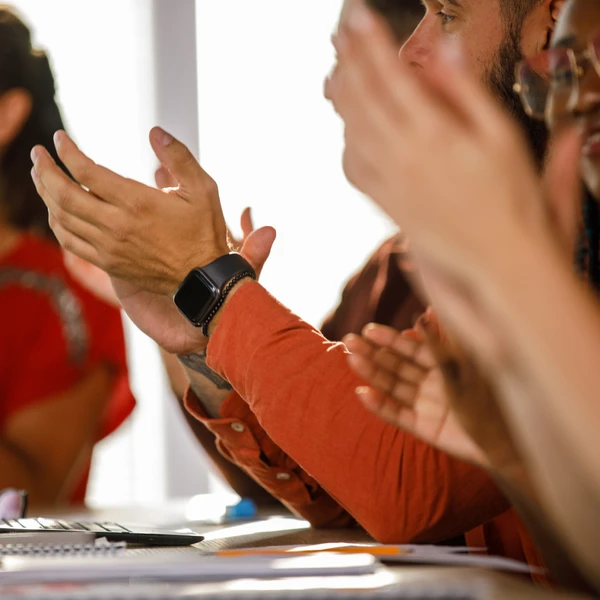 People sitting at a table, clapping. A person in the foreground wears a smartwatch. Notebooks and a laptop are visible on the table.