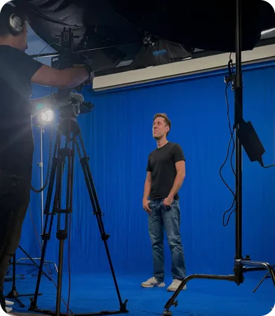 A person in a black t-shirt and jeans stands in front of a blue screen backdrop in a professional video production studio, being filmed by a camera operator in the foreground with studio lighting rigs overhead.