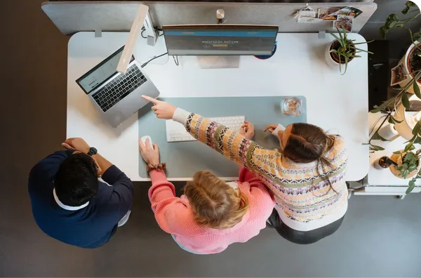 Three people collaborating at a desk with a laptop and monitor. One person points at the screen. Plants and office items are visible.