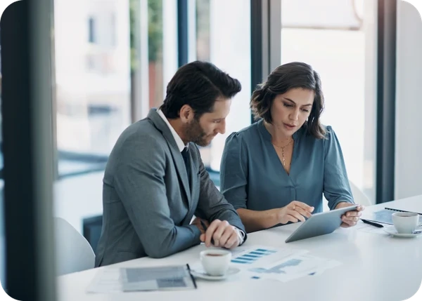 Two colleagues reviewing a tablet and charts at a bright office table with coffee cups.