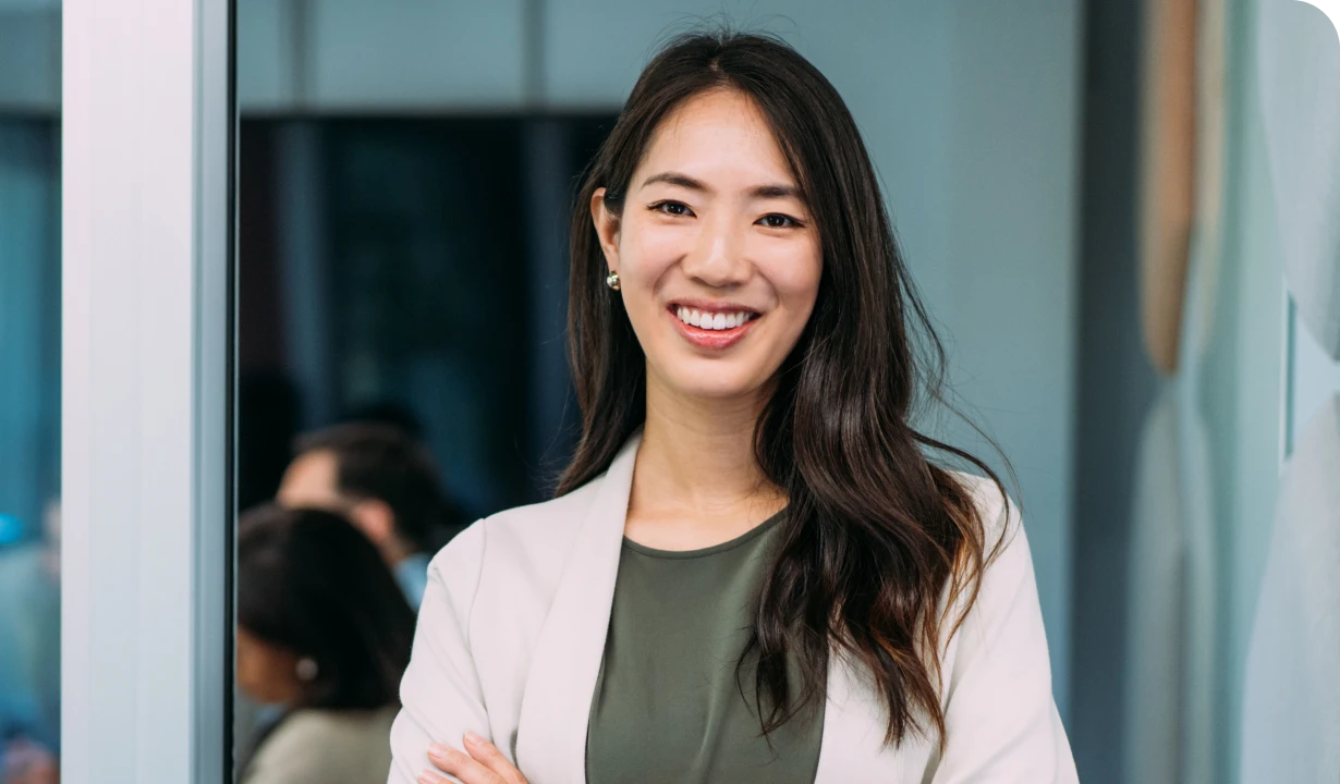 Smiling woman with long hair in a white blazer and green top, standing confidently with arms crossed in an office setting.