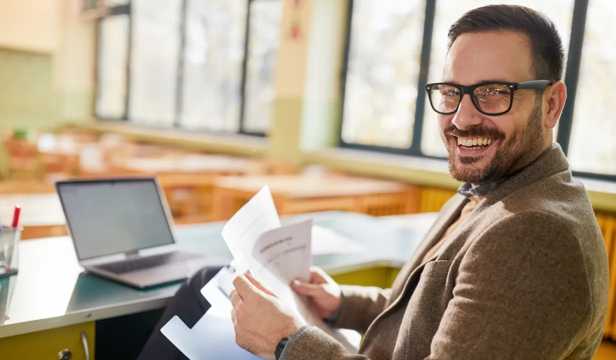 Smiling man with glasses holding papers, sitting at a desk with a laptop in a bright room with large windows.