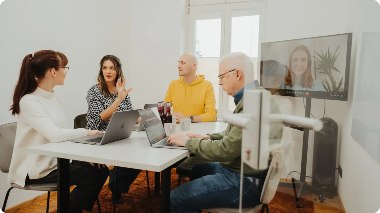 Four Staffbase colleagues in a meeting room with laptops, joined by a remote colleague on a video screen.