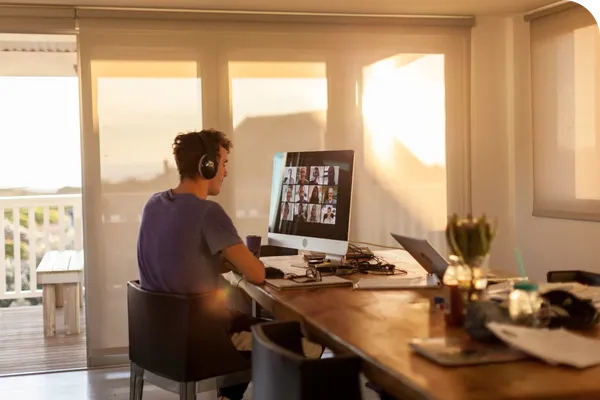 Person wearing headphones, sitting at a desk in a sunlit room, participating in a video conference on a computer screen.