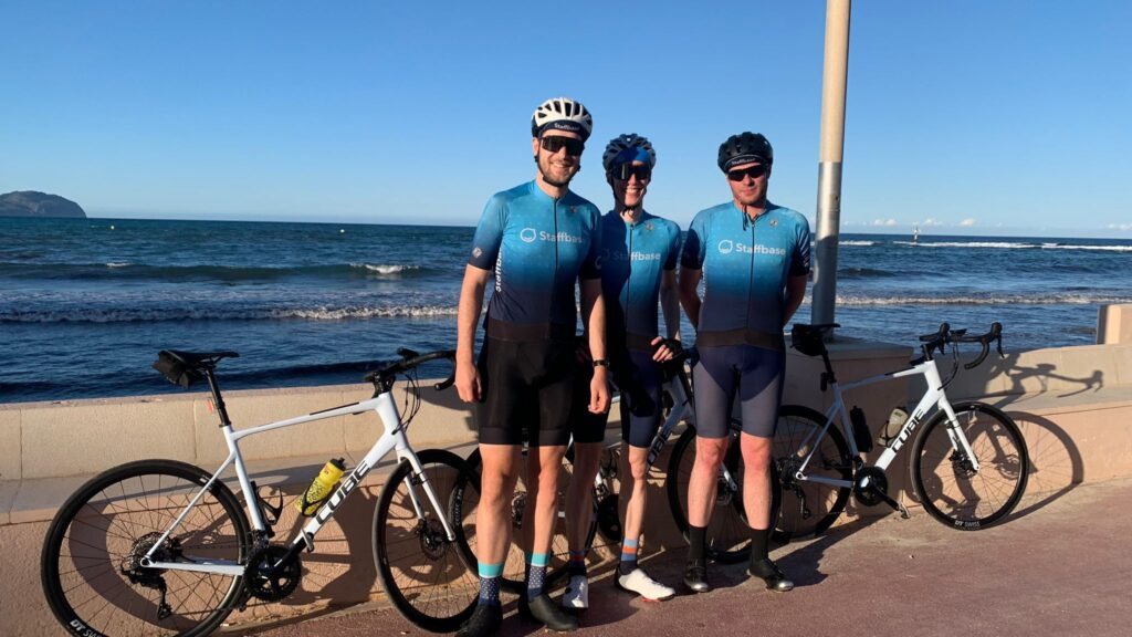 Three cyclists in matching blue Staffbase jerseys and helmets stand smiling beside white Cube road bikes on a seaside promenade.