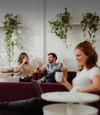 Staffbase colleagues relaxing on a burgundy sofa with coffee mugs, chatting under hanging plants.