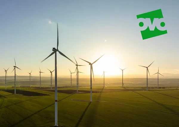 Wind turbines on a grassy field at sunset, with the sun low on the horizon and a green logo in the sky.