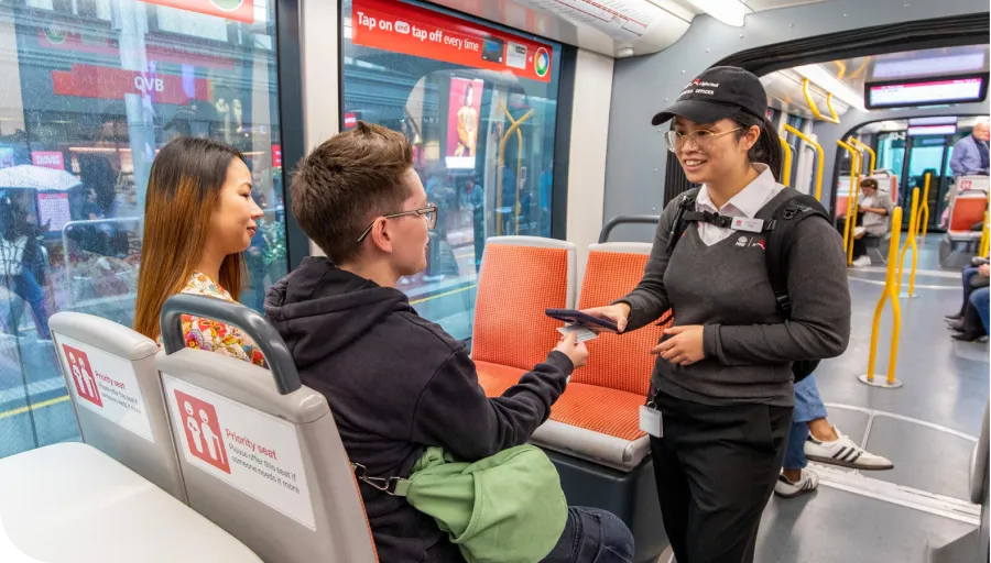 A transit officer checks a passenger's ticket on a tram, while another passenger sits nearby. The tram interior is modern and well-lit.