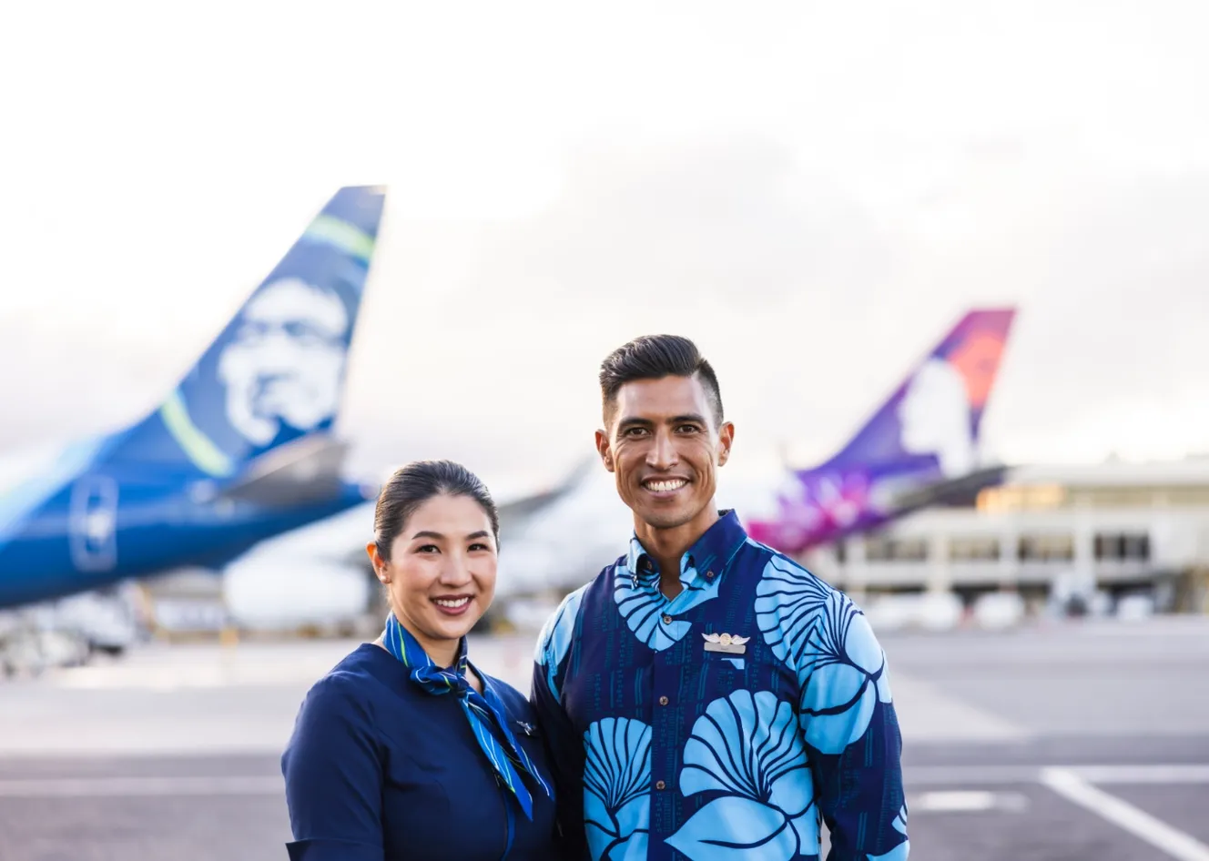 Two airline crew members in blue uniforms smile on an airport tarmac, with airplanes in the blurred background.