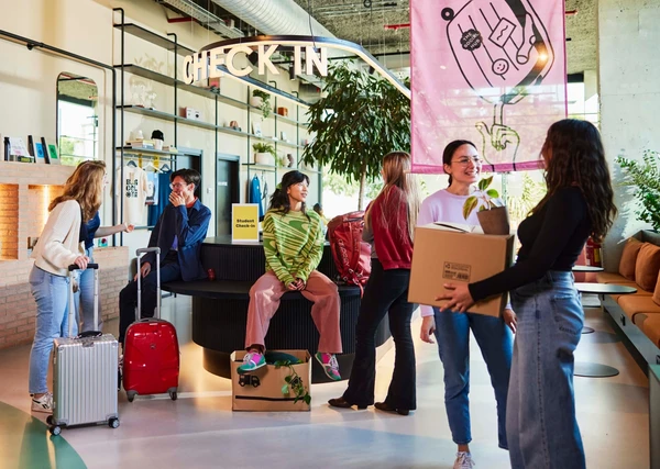 People with luggage and boxes gather in a modern check-in area, chatting and smiling. A large "CHECK IN" sign is visible above.