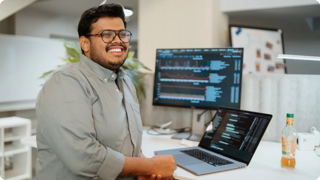 Staffbase engineer smiling at his desk with a code editor open on his laptop and a data dashboard on his monitor.