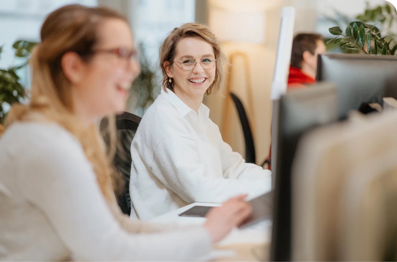Two people in an office, one smiling at the camera, both seated at desks with computers, surrounded by a bright and modern workspace.