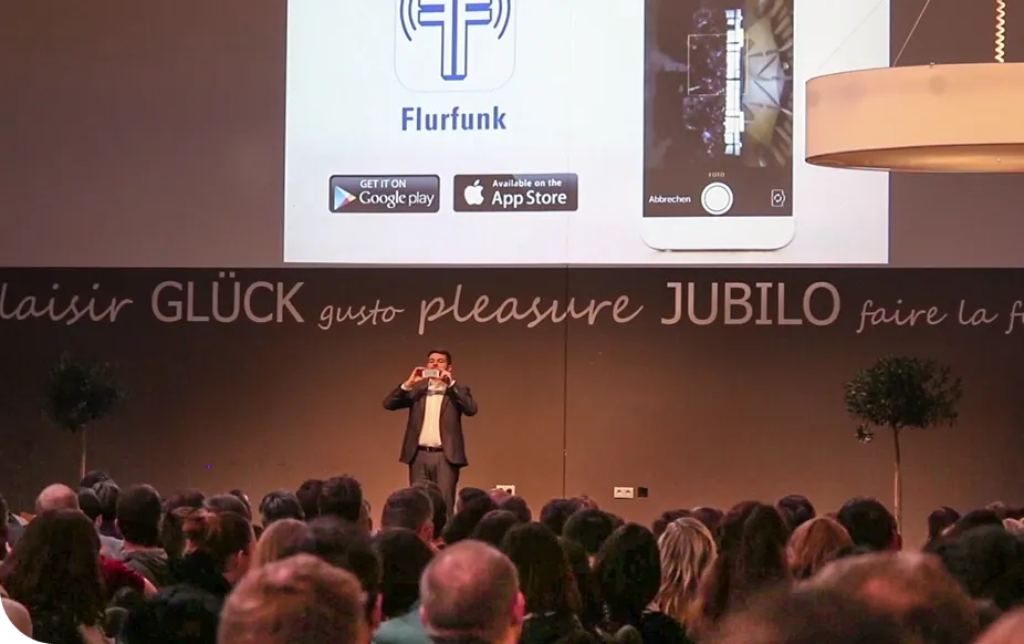 A speaker presents on stage to a large audience, with a screen displaying a smartphone app and download options from Google Play and the App Store.