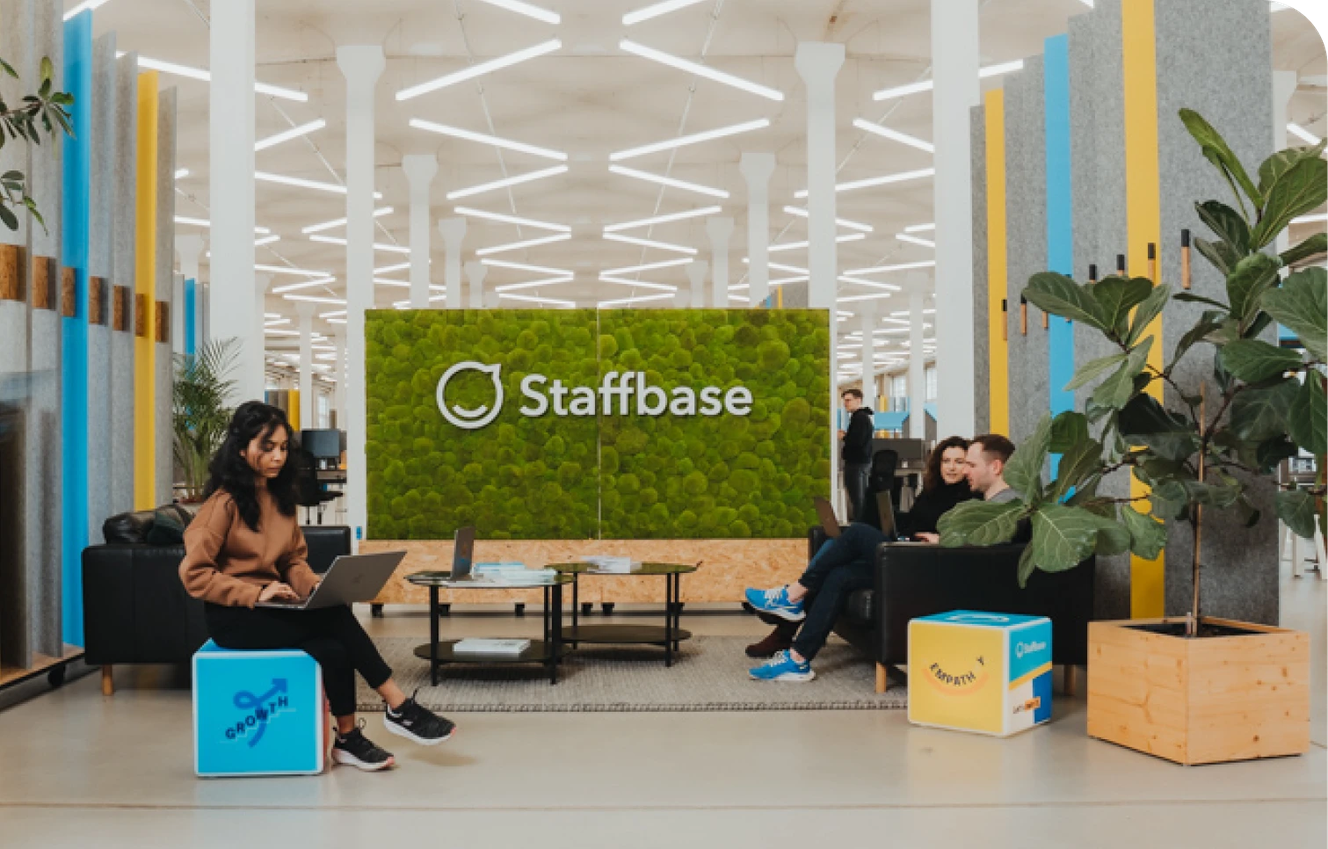Modern office space with people sitting on sofas, working on laptops. A green wall displays the "Staffbase" logo. Bright, open environment.