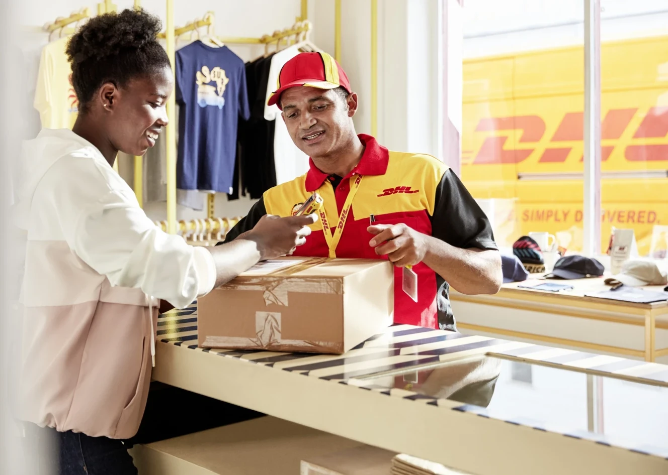 A customer hands a package to a DHL employee at a counter. The employee wears a DHL uniform and cap. Clothing items are displayed in the background.