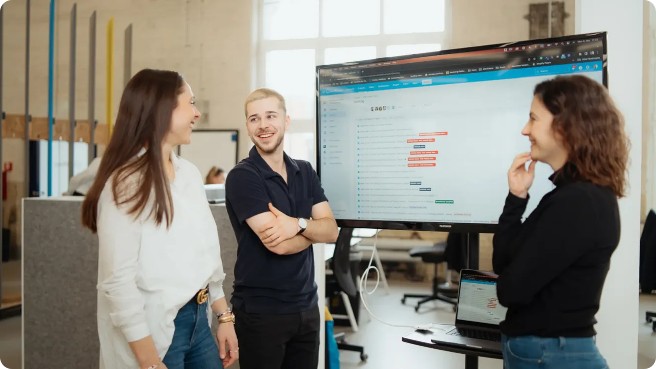 Three Staffbase colleagues chatting in front of a large screen displaying a project management dashboard.