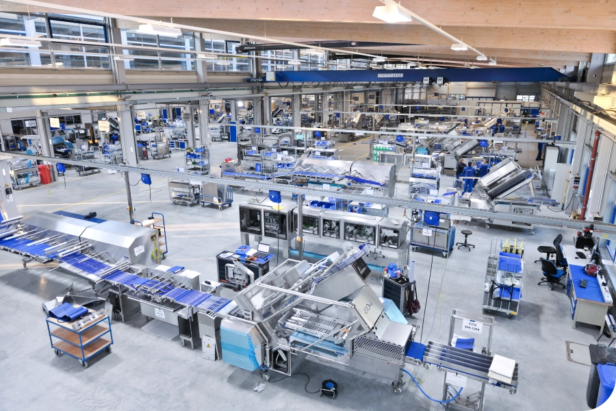 A large, modern factory interior with automated machinery and conveyor belts, featuring blue accents and organized workstations.