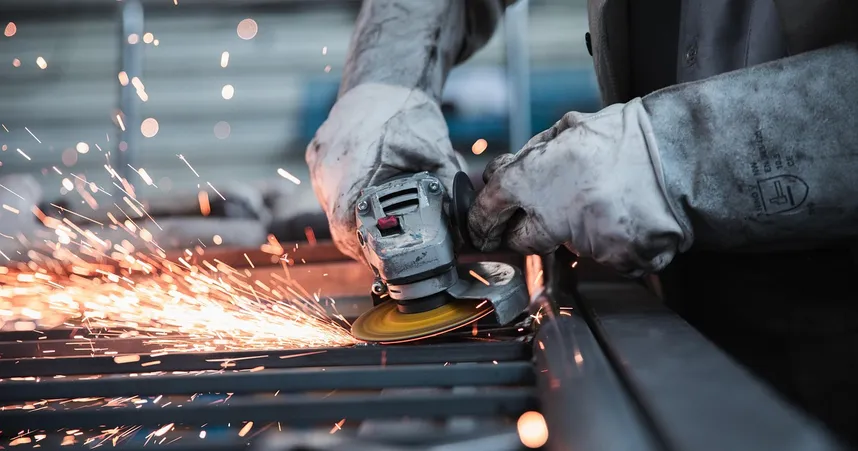 Worker using an angle grinder, creating sparks on a metal surface in an industrial setting. Wearing protective gloves.