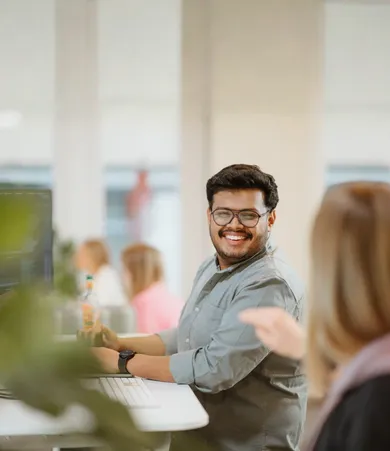Staffbase employee smiling at his desk while chatting with a colleague in an open-plan office.