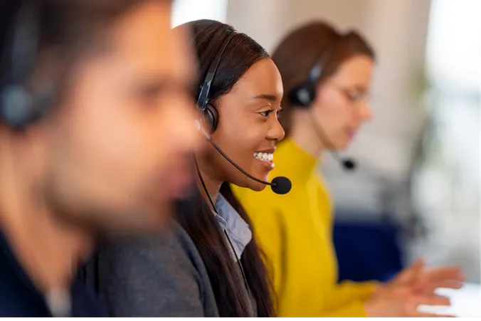 Three customer service representatives wearing headsets, focused and smiling, working at desks in a bright office environment.