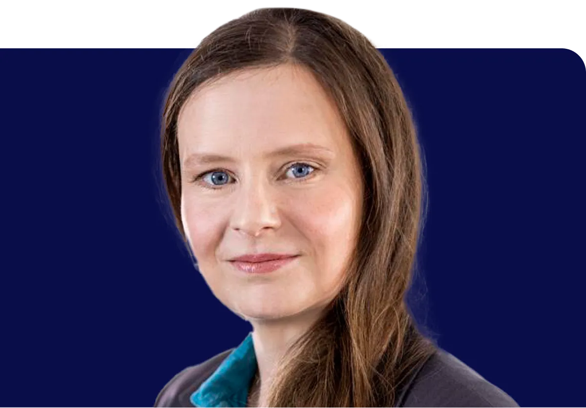 Woman with long brown hair, wearing a blue collared shirt and dark blazer, smiling against a dark blue background.