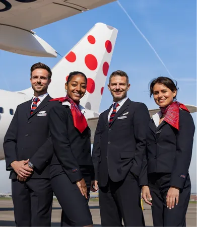 Four airline crew members in uniform stand smiling in front of an airplane with a polka-dotted tail under a clear blue sky.