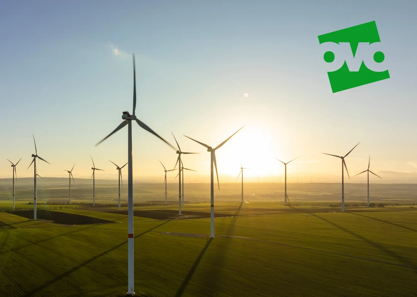 Wind turbines on a grassy field at sunset, with the sun low on the horizon and a green logo in the sky.