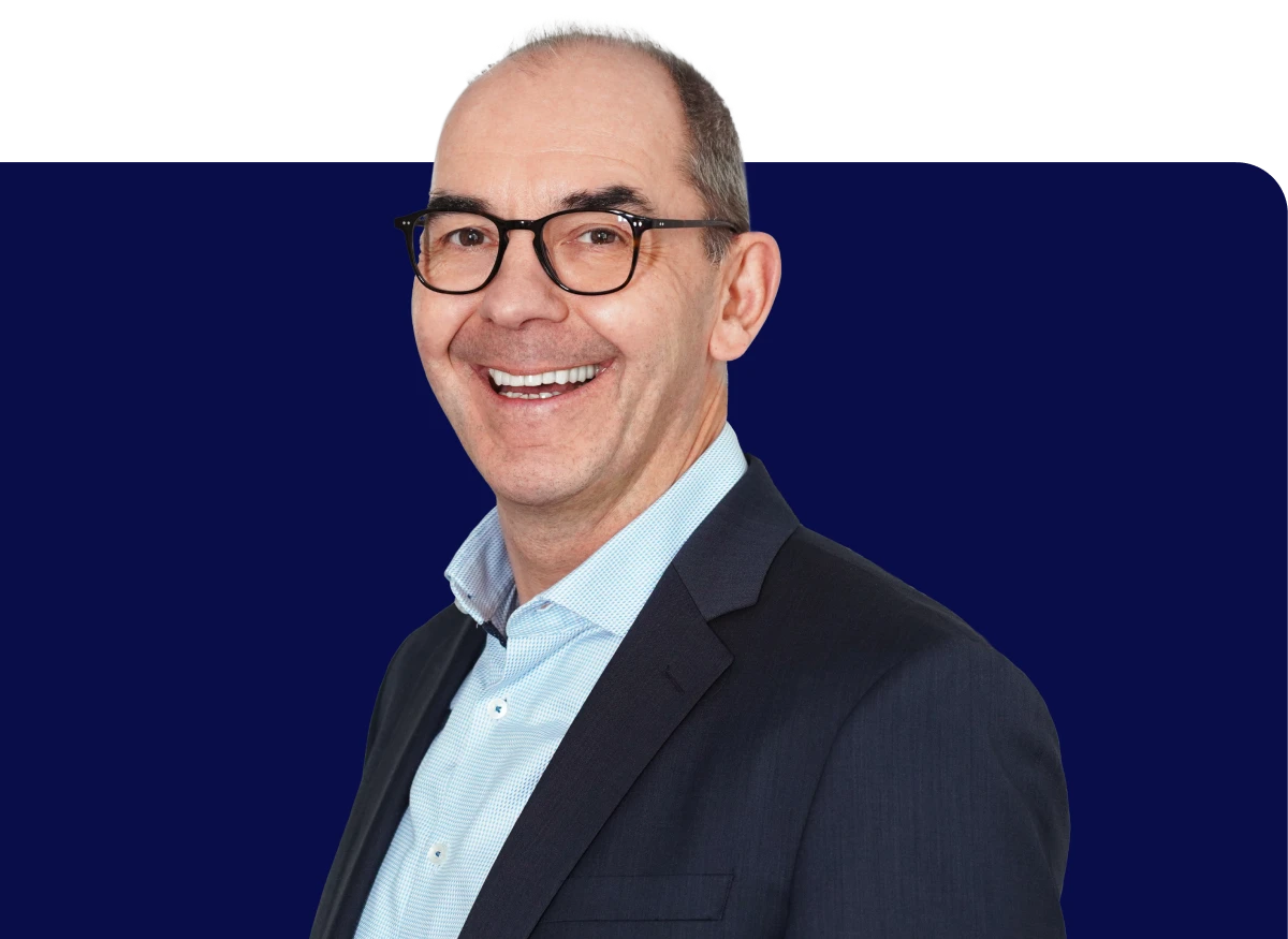 Smiling man in glasses and a dark suit with a light blue shirt, standing against a dark blue background.