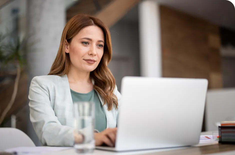 Woman with long hair working on a laptop at a desk, wearing a light blazer. A glass of water is on the table in a modern office setting.