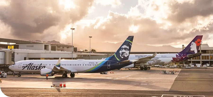 Two airplanes parked at an airport gate during sunset, with Alaska Airlines and another airline featuring a floral tail design.