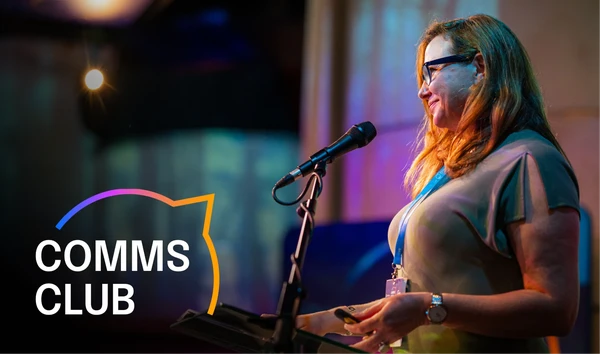 Woman speaking at a podium with a microphone, wearing glasses. "COMMS CLUB" text with colorful outline is visible on the left.
