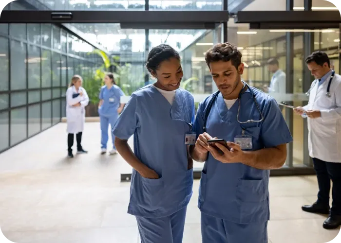 Two medical staff in blue scrubs looking at a smartphone in a hospital hallway, colleagues conversing in the background.