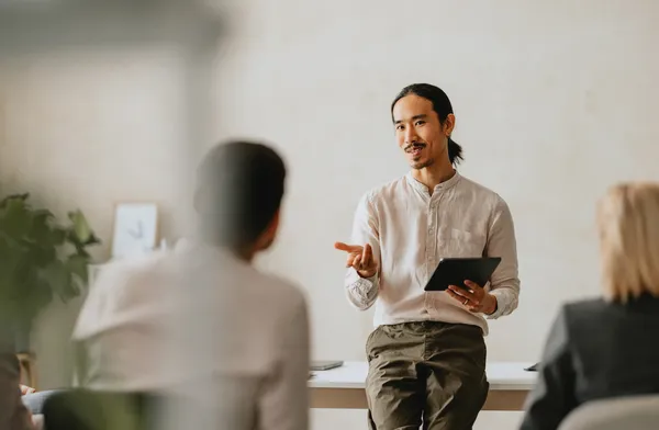 A person sitting on a table, holding a tablet, and speaking to a small group in a bright room.