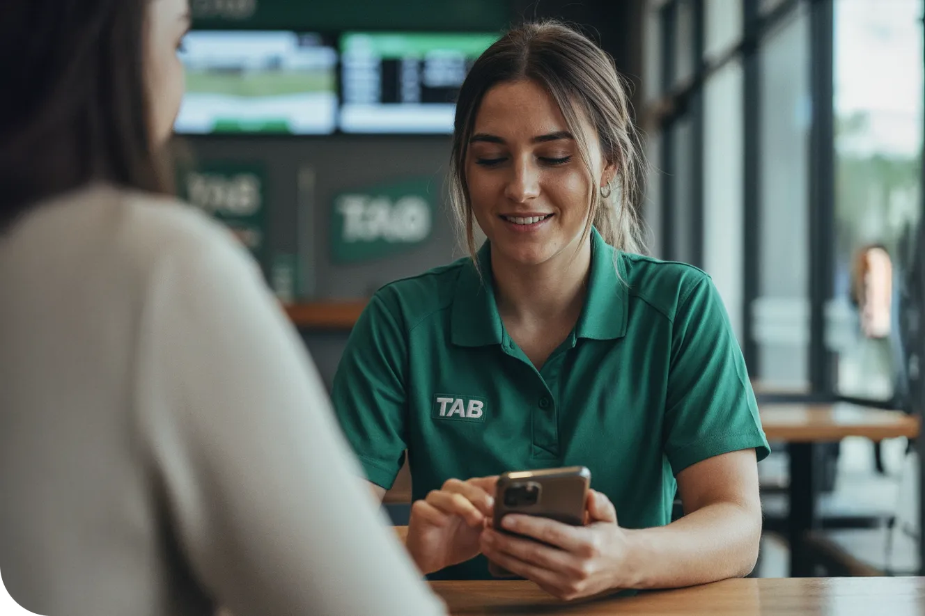 A close-up of a smiling woman wearing a green TAB uniform sitting at a wooden table and looking at her smartphone.