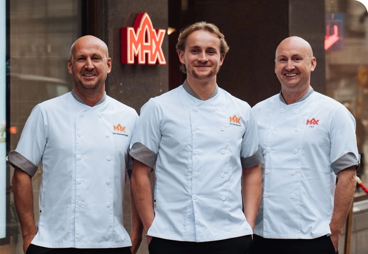 Three smiling chefs in white uniforms stand in front of a building with a red "MAX" sign.