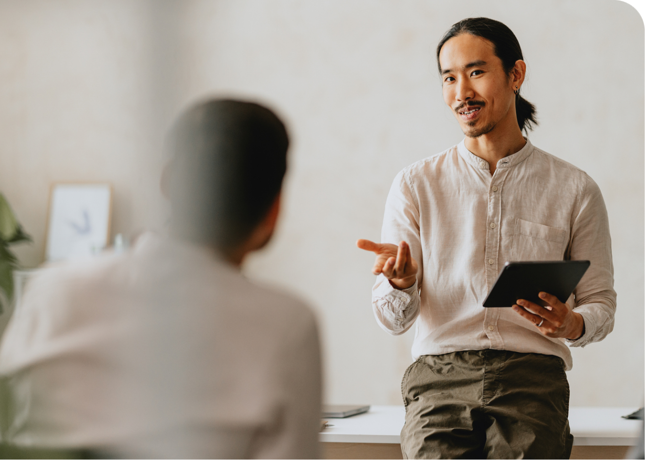 Man with a tablet gestures while sitting on a desk, engaging in conversation with another person in a blurred foreground.