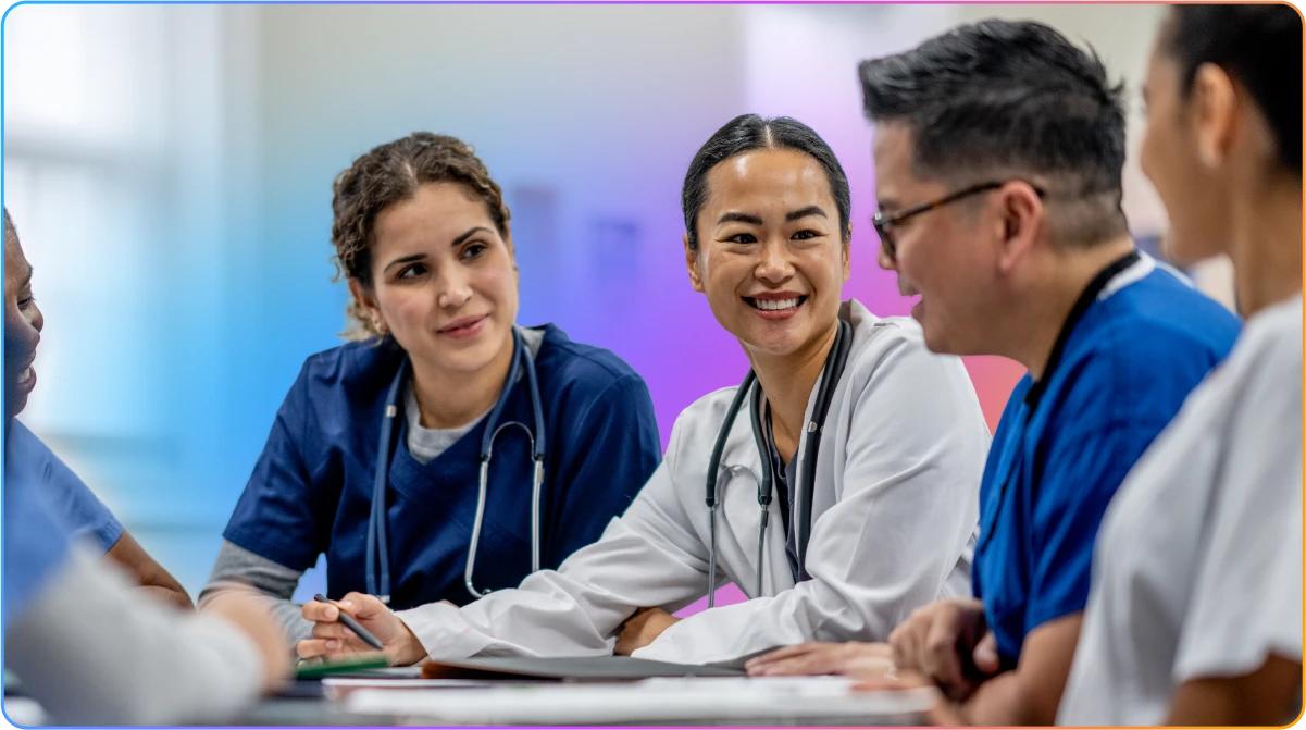 A diverse group of smiling healthcare professionals in scrubs and lab coats having a discussion around a table.