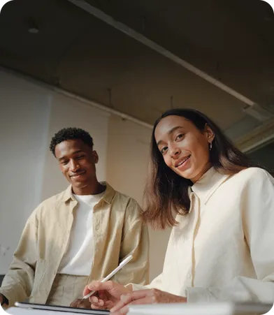 Two people smiling while working together at a table, holding a tablet and pen in a bright room.