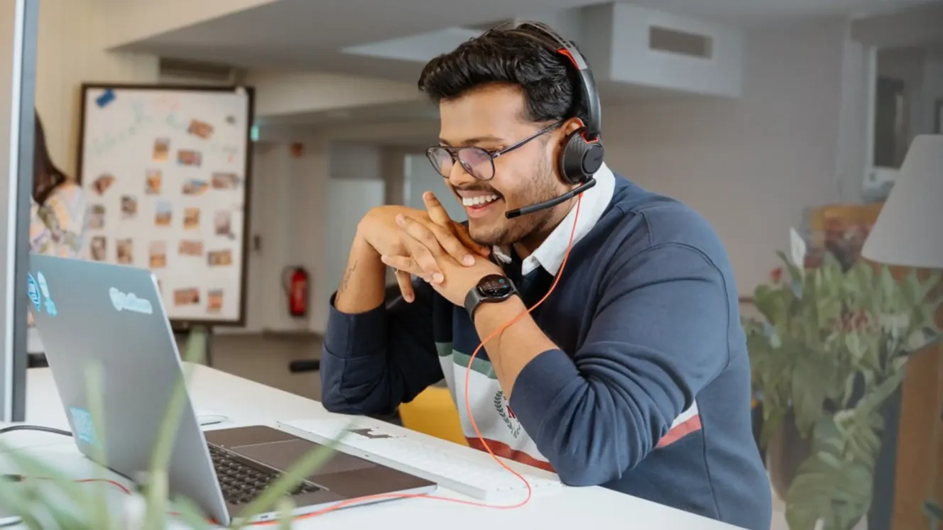 Staffbase employee smiling during a video call, wearing a headset at his laptop in the office.