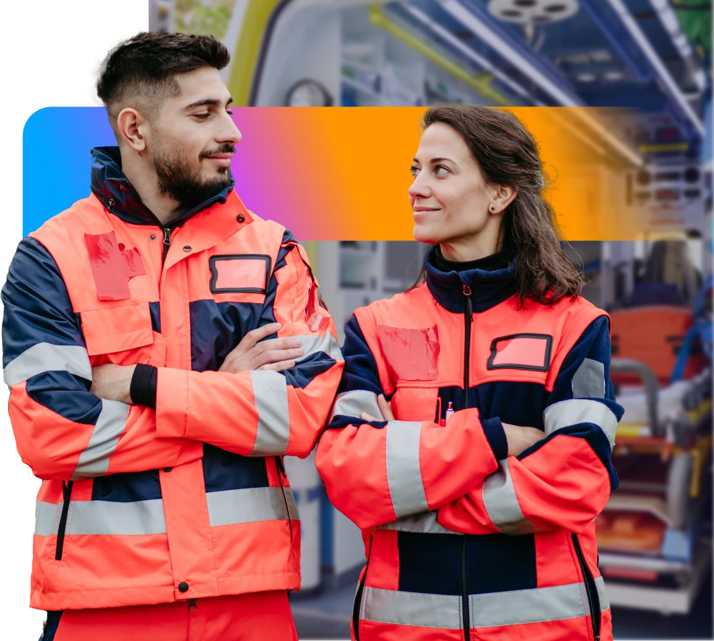 Two people in bright orange and navy uniforms stand with arms crossed, smiling at each other, in front of an ambulance interior.